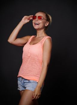 Cheerful smiling woman with tanned skin, beautiful muscular fit body in denim shorts, looking through her pink sunglasses and showing tongue to the camera, posing over black background copy space