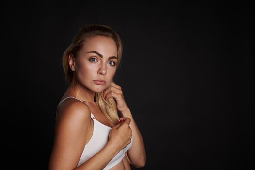 Confident portrait of an attractive young Caucasian woman with perfect clean skin and beautiful blond hair looking at camera standing against black wall background with copy space