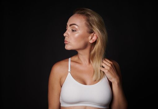 Confident portrait of a beautiful blonde middle aged woman in white underclothing looking aside posing over black background with copy ad space