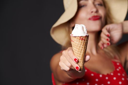 Soft focus on ice cream in the hands of a cheerful sexy blonde woman in red swimsuit with white polka dots and a summer hat, isolated over black background with copy ad space. Summer holidays concept