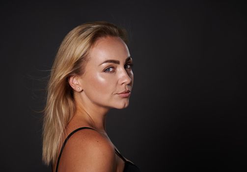 Studio shot of confident blonde beautiful middle aged woman with healthy tanned clean glowing skin in black bra looking at camera standing three quarters on dark background with copy space. Close-up