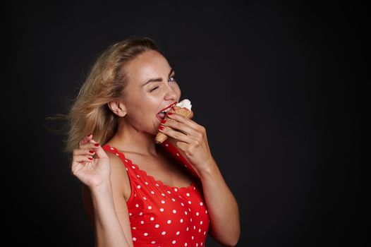 Attractive stunning blonde European woman with flying hair from the wind, wearing a red swimsuit with white polka dots eats ice cream cone, looks at camera posing against black background, copy space