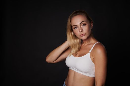 Beautiful young blonde woman with perfect aesthetic body in white sport top confidently looking at camera, isolated over back background with copy ad space