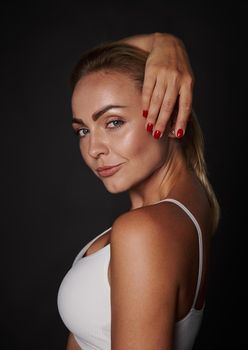 Side portrait of a beautiful attractive confident tanned young Caucasian woman with straight blond hair, natural make-up, beautiful healthy glowing skin posing at the camera on a dark background