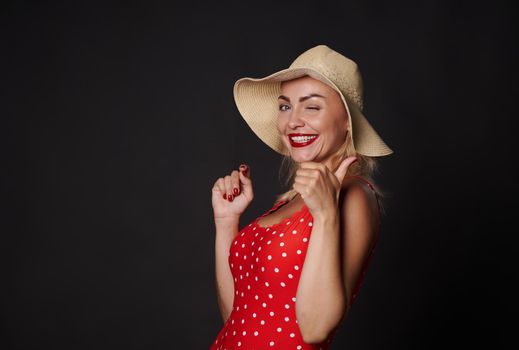Attractive stunning blonde woman in summer straw hat and red swimsuit winking and smiling with beautiful white toothy smile, showing thumb up at camera, posing against black background with copy space