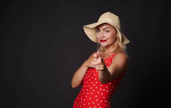 Attractive blonde European woman in red outfit with white polka dots and straw summer hat smiles beautiful white toothy smile, pointing up finger at copy space on black background