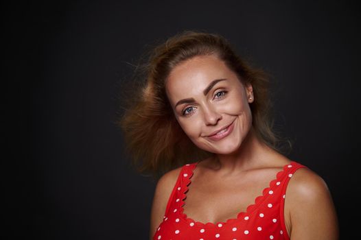 Headshot of stunning beautiful Caucasian woman with fresh glowing skin, natural make-up and flying shiny blond hair in red top smiling toothy smile looking at camera isolated over black background