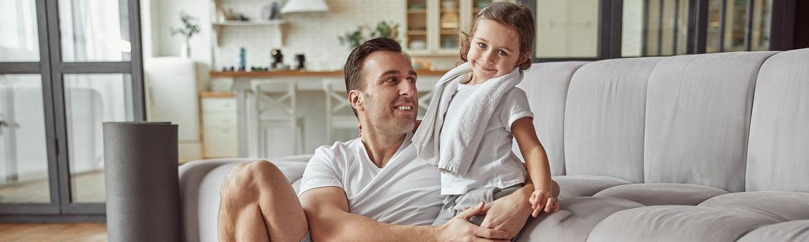 Full length portrait of smiling girl in dad hugs in living room while man is doing workout indoors