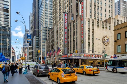 Radio City Music Hall, New York City with two yellow Taxi Cabs and traffic in the forefront, people walking on the sidewalk during daytime, horizontal
