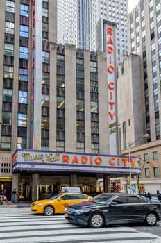 Radio City Music Hall New York City with crosswalk and traffic, a yellow taxi and black car in the forefront during daytime, vertical