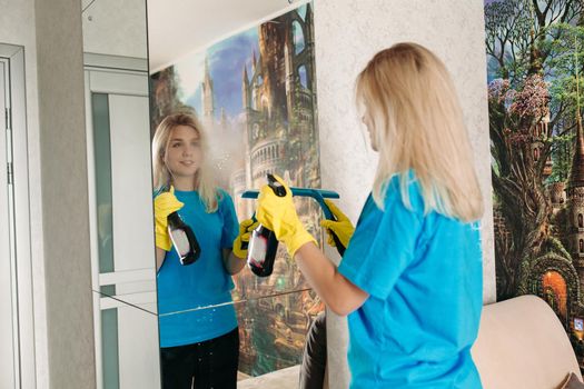 Young girl wearing in yellow protective rubber gloves, with special detergent spray and brush, washing mirror in room. Female employee of cleaning company wearing in uniform working at client home.