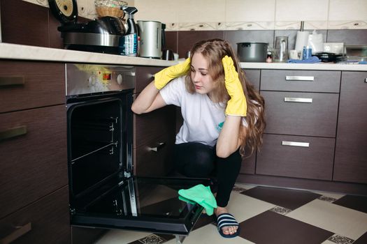 Young woman in yellow protective gloves holding head by hands, looking dirty oven at home kitchen. Brunette girl with curly hair washing oven on kitchen holding hand inside. Housework concept.