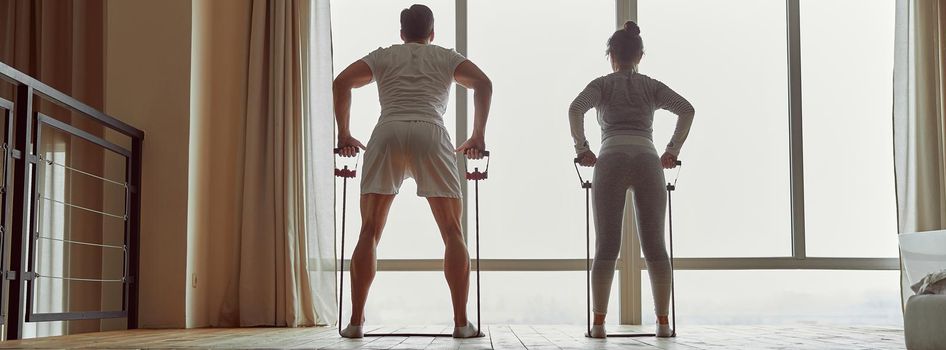 Sporty man is doing workout with wife and using resistance bands in front of window