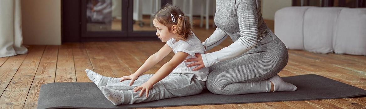 Happy mother is enjoying stretching exercises together with little girl and assisting her in living room