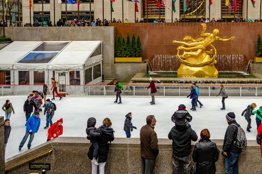 The Rink At Rockefeller Center during winter, Lots of People skating on ice while people standing in front watching them, golden Prometheus statue and flags in the background, horizontal, horizontal