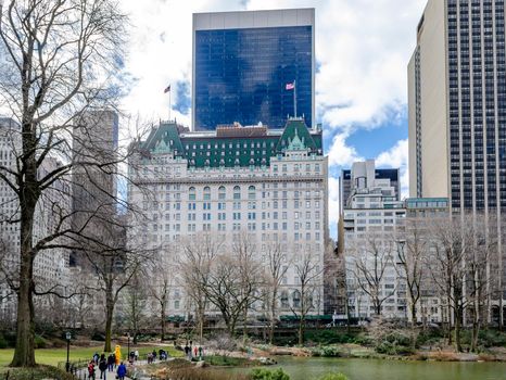 The Pond at Central Park Lake during winter with The Plaza Hotel in Background during daytime, People walking next to the Lake, water and trees in forefront, horizontal