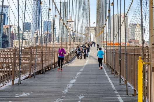 Two women jogging on Brooklyn Bridge New York City during daytime, lots of tourist walking in the background, wire ropes in the forefront, skyline of Manhattan in the Background, Horizontal