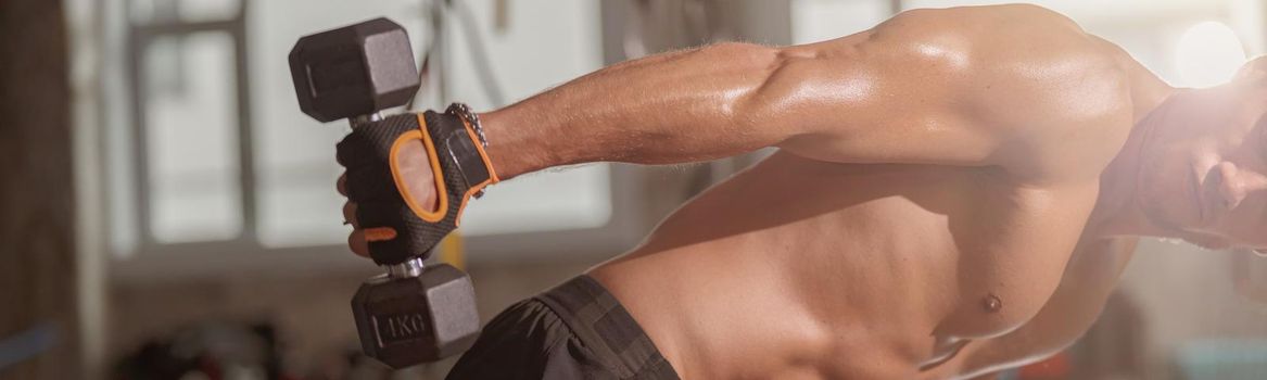 Athletic man in workout gloves standing, holding black dumbbell, pulling it to the side and bending down doing hand exercise