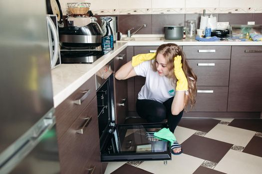Young woman in yellow protective gloves holding head by hands, looking dirty oven at home kitchen. Brunette girl with curly hair washing oven on kitchen holding hand inside. Housework concept.