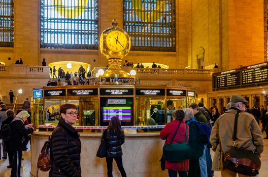 Information Building in Grand Central Terminal indoors with people waiting in forefront,  golden colored yellow Clock above the information, lots of people on the first floor of the building, staircase and window in background, horizontal