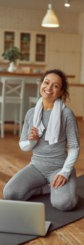 Full length portrait of smiling female with towel sitting on mat during training with notebook at home