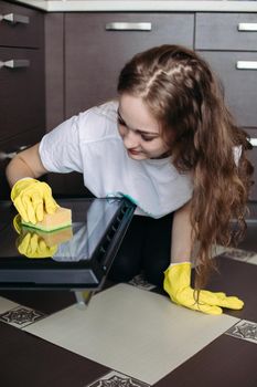 Close up of woman, wearing in protective glove with rag cleaning oven at home kitchen. Beautiful brunette girl, with curly hair on her knees washing oven on kitchen in brown color. Housework concept.
