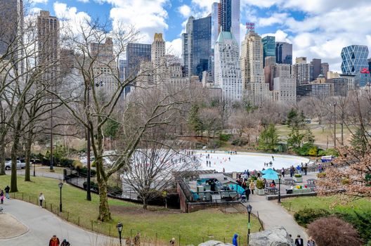 Trumps Wollman Rink with People Ice Skating during daytime in winter, view from the distance, Central Park New York City, benches and people walking in the forefront, horizontal