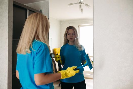 Female employee of cleaning company wearing in uniform working at client home. Blonde girl wearing in yellow protective rubber gloves, with special detergent spray and brush, washing mirror.