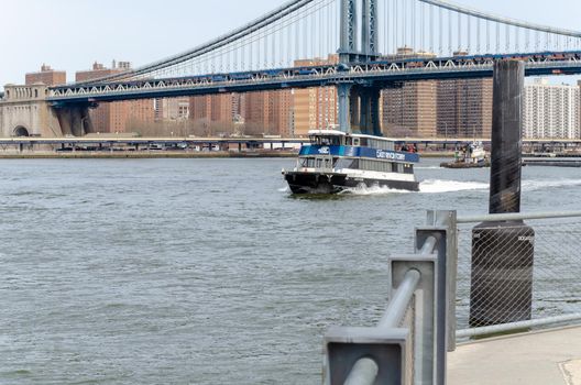 Blue East River Ferry underneath of Manhattan Bridge, lots of tourist with jackets standing on top of the ship, New York City, during winter, with steel fence in forefront, horizontal