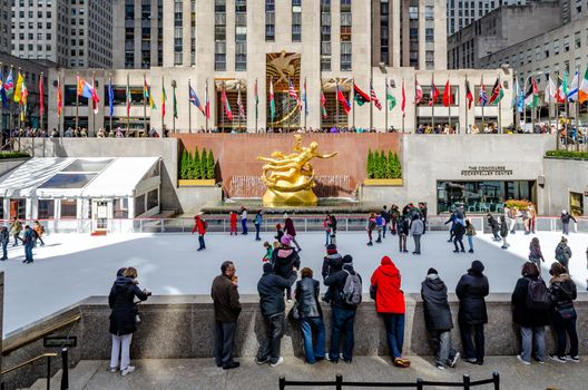 The Rink At Rockefeller Center during winter, Lots of People skating on ice while people standing in front watching them, golden Prometheus statue and flags in the background, horizontal