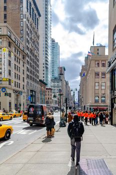 Lots of People standing and walking on a sidewalk in Manhattan, New York City, Promoter with orange sweatshirt talking to People, Skyscraper and yellow taxi cabs on the street, vertical