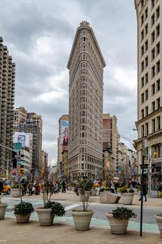 Flatiron Building New York City during daytime with overcast, lots People, traffic, yellow taxi cabs and Plants in forefront, view from low angle, vertical