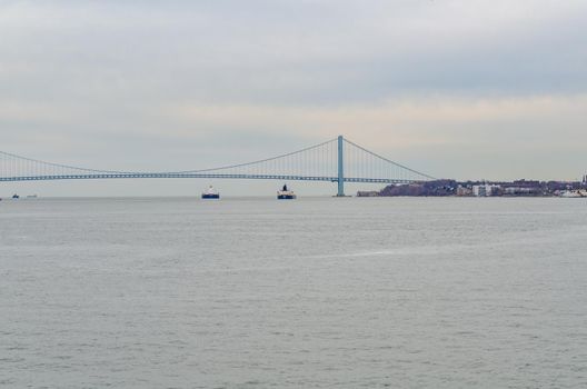 Two industrial ships underneath blue Verrazano-Narrows Bridge, New York City, at the horizon, during winter evening with overcast, horizontal