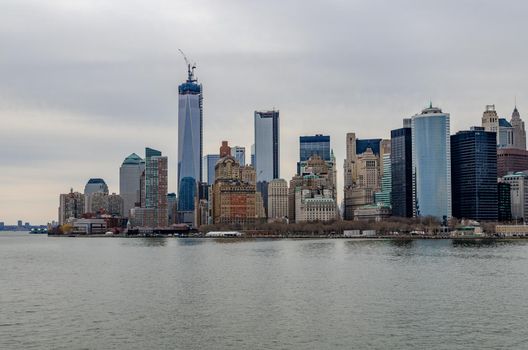 Skyline of Manhattan with One World Trade Center with construction area and Crane on top of it and Hudson River in forefront, during winter evening with overcast, horizontal