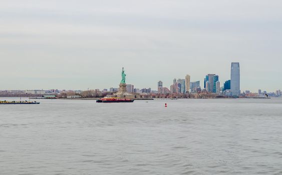 Statue of Liberty National Park side view, New York City with industrial ship passing by in the forefront, during winter day with overcast, horizontal