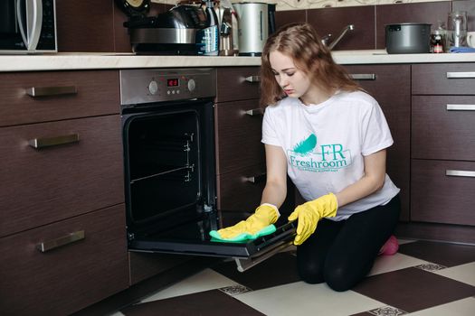 Young woman in yellow protective gloves holding head by hands, looking dirty oven at home kitchen. Brunette girl with curly hair washing oven on kitchen holding hand inside. Housework concept.