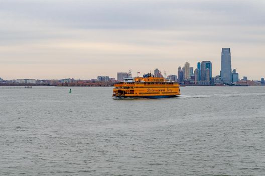 Yellow Staten Island Ferry on Hudson river passing by in front of Jersey City with office building skyscraper, New York, during winter evening with overcast, horizontal