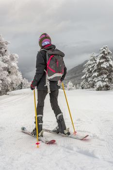 Woman with black ski wear, helmet and backpack skiing down the top of the mountain in ski centre, stryn with clouds and trees in the background, rear view, vertical