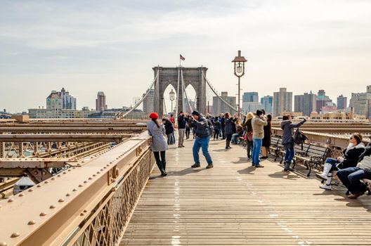 Brooklyn Bridge New York City in the evening during winter with tourists walking, taking photos and relaxing on benches on the bridge, wide angle shot, skyscraper in the background, horizontal