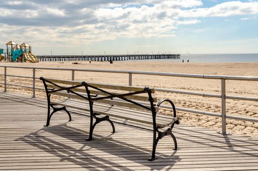 Bench at Beach Promenade of Coney island, rear view, with Pat Auletta Steeplechase Pier in Background, Brooklyn New York City during sunny winter day with cloudy sky, Playground on the Beach, horizontal