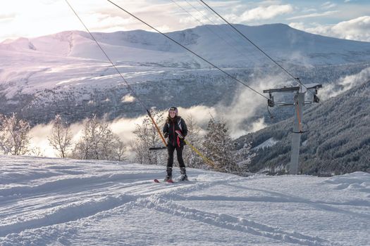 Woman with brown hair smiling, wearing black ski wear, on ski lift moving up, beautiful mountaing range and valley in background with sunlight, horizontal