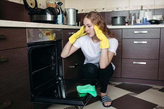 Young woman in yellow protective gloves holding head by hands, looking dirty oven at home kitchen. Brunette girl with curly hair washing oven on kitchen holding hand inside. Housework concept.