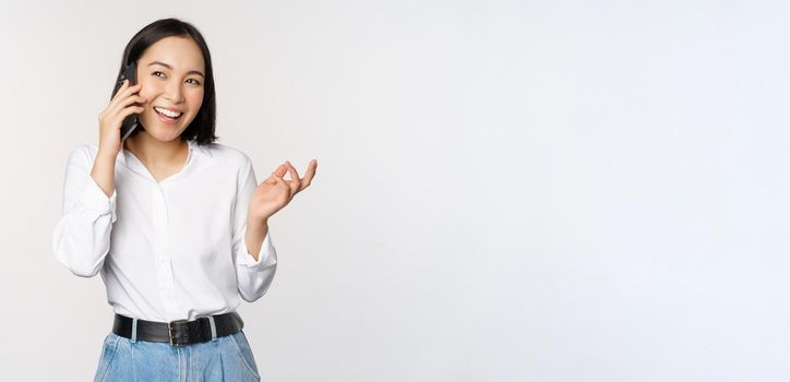 Friendly smiling asian woman talking on phone, girl on call, holding smartphone and laughing, speaking, standing over white background.