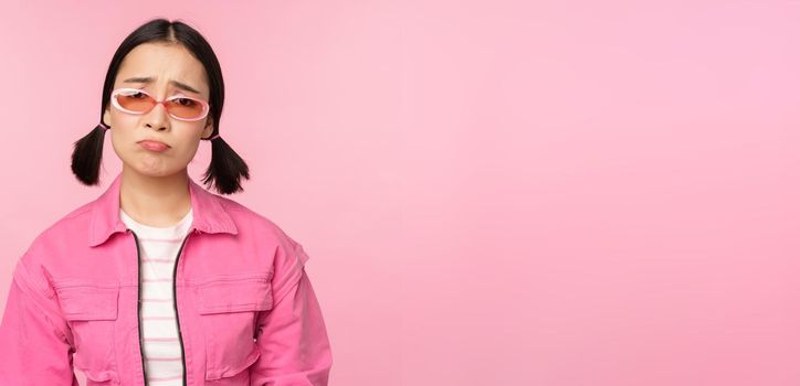 Portrait of sad and gloomy asian girl sulking from disappointment, standing upset against pink studio background. Copy space