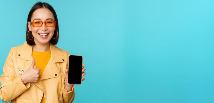 Young stylish chinese woman showing mobile phone screen and thumbs up, smartphone app, standing over blue background. copy space