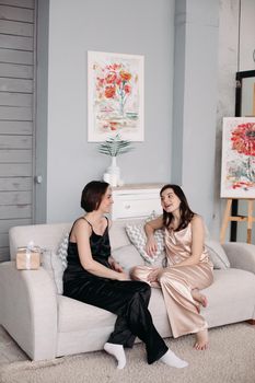 Side view of two young sisters sitting on sofa at home and talking. Attractive girls in pajamas looking at each other, laughing and relaxing in morning. Concept of rest and weekends.