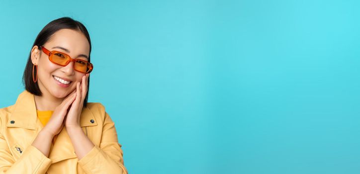 Close up portrait of asian young woman in sunglasses, smiling and looking romantic, standing happy over blue background. Copy space