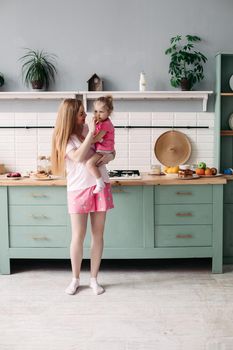 Full length of beautiful smiling woman in pyjamas holding her lovely daughter on hands and feeding her with fruit for breakfast. Modern kitchen.