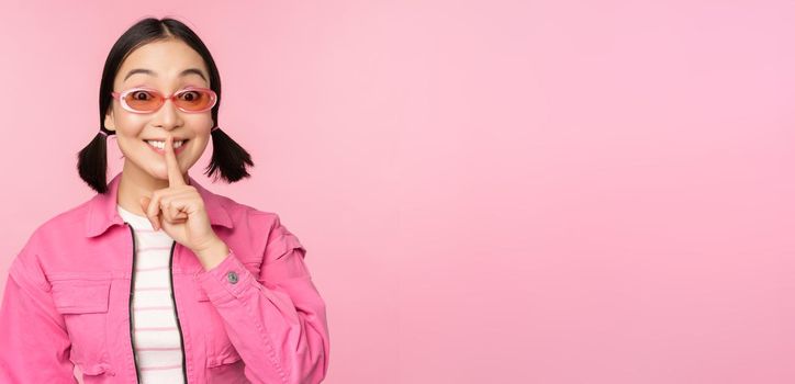 Modern korean girl in stylish spring outfit, sunglasses, showing shush, hush sign, press finger to lips, taboo gesture, standing over pink background.