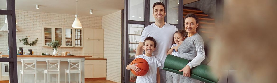Full length portrait of joyful parents with son and daughter standing with sport equipment in living room